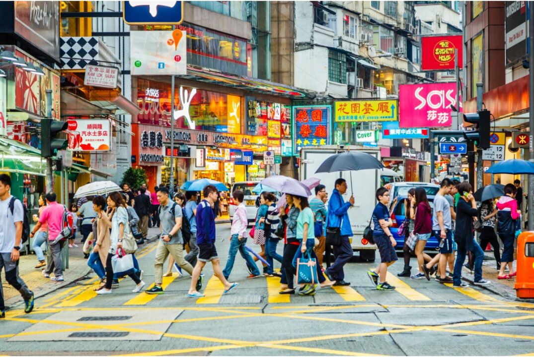 People cross a busy street in a shopping district in Hong Kong. Many of them hold umbrellas and shopping bags.