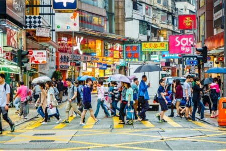 More Than 100,000 Hong Kong Residents Returned To City In 2022-2023, HK Population Will Cross 8 Million In 2046 People cross a busy street in a shopping district in Hong Kong. Many of them hold umbrellas and shopping bags.