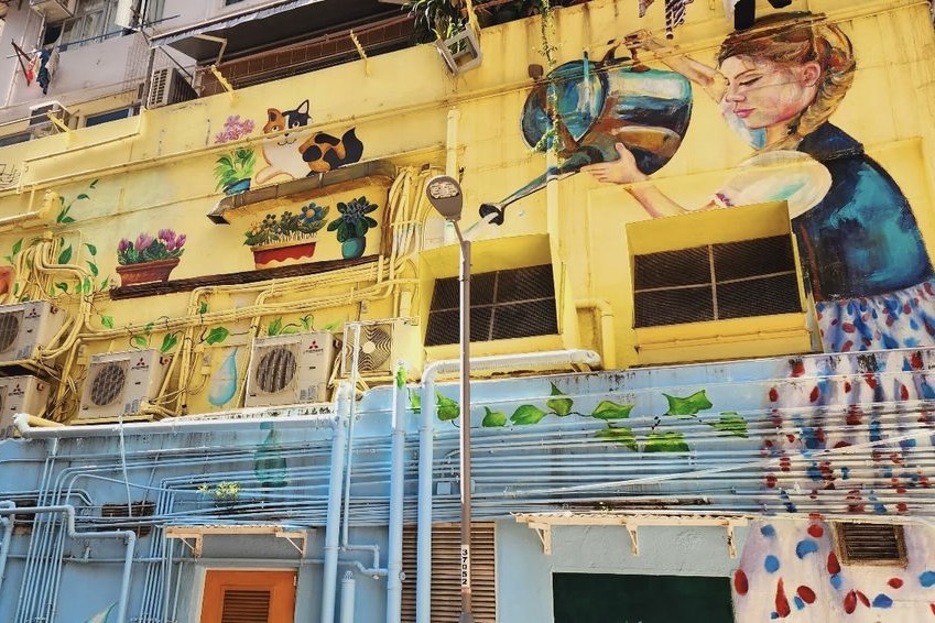 A mural depicting a girl watering potted plants. She is wearing a blue-and-white short-sleeved blouse with a long patterned skirt and uses a tin watering can. There is a tabby cat in the mural, as well as creeping plants at the bottom.