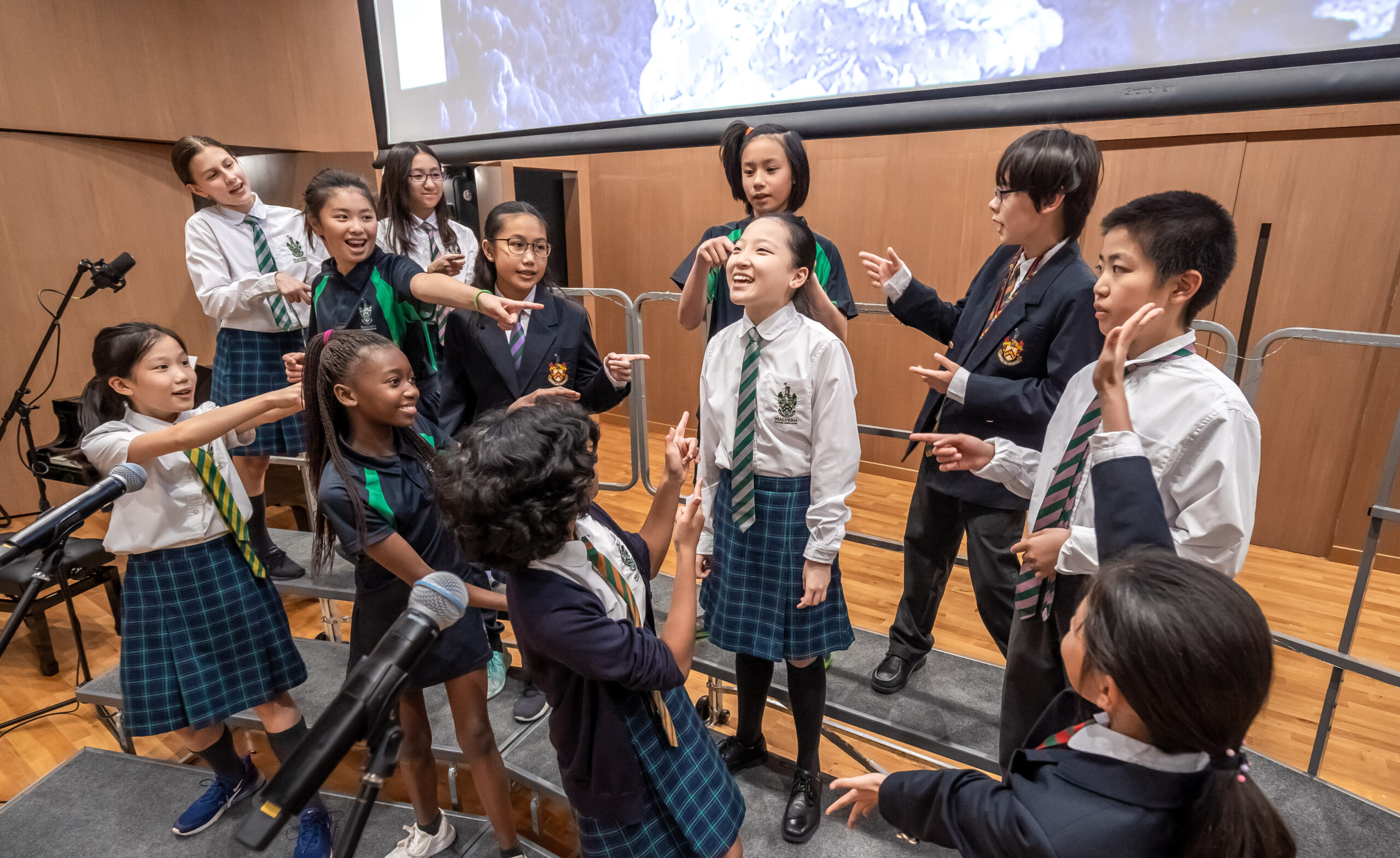 Students practise a musical number at Malvern College Hong Kong. One student, a girl, stands at the centre of a group while other children point at her. The children all wear the Malvern uniform, and they are either smiling or singing. 