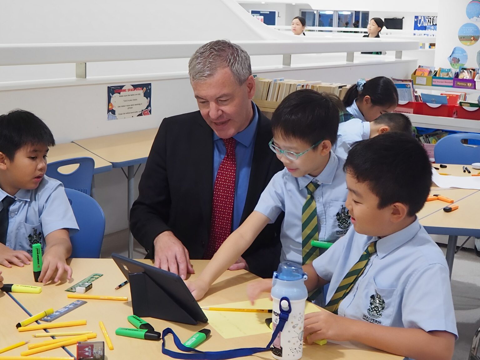 The principal of Malvern College Hong Kong sits among young students in a classroom. They are all looking at a tablet. The table at which they are seated has pencils, highlighters, rulers, and other stationery on it.