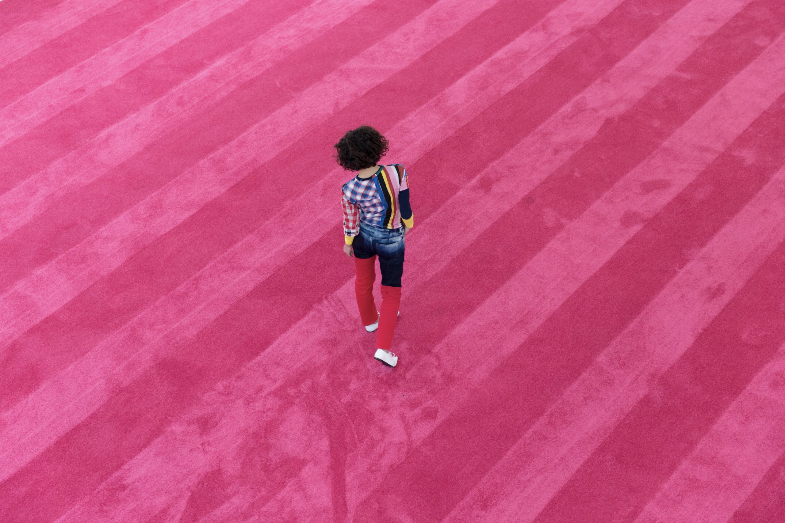 A performer wearing multi-coloured clothing in a variety of patterns and fabrics -- plaid, stripes, and denim -- walks across a striped pink carpet.