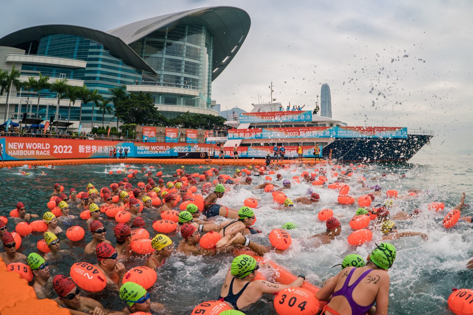 Swimmers at the beginning of the New World Harbour Race in Hong Kong. They all wear orange or green swim caps and are swimming towards the Tsim Sha Tsui finish point across the harbour. The Hong Kong Convention and Exhibition Centre is in the background.