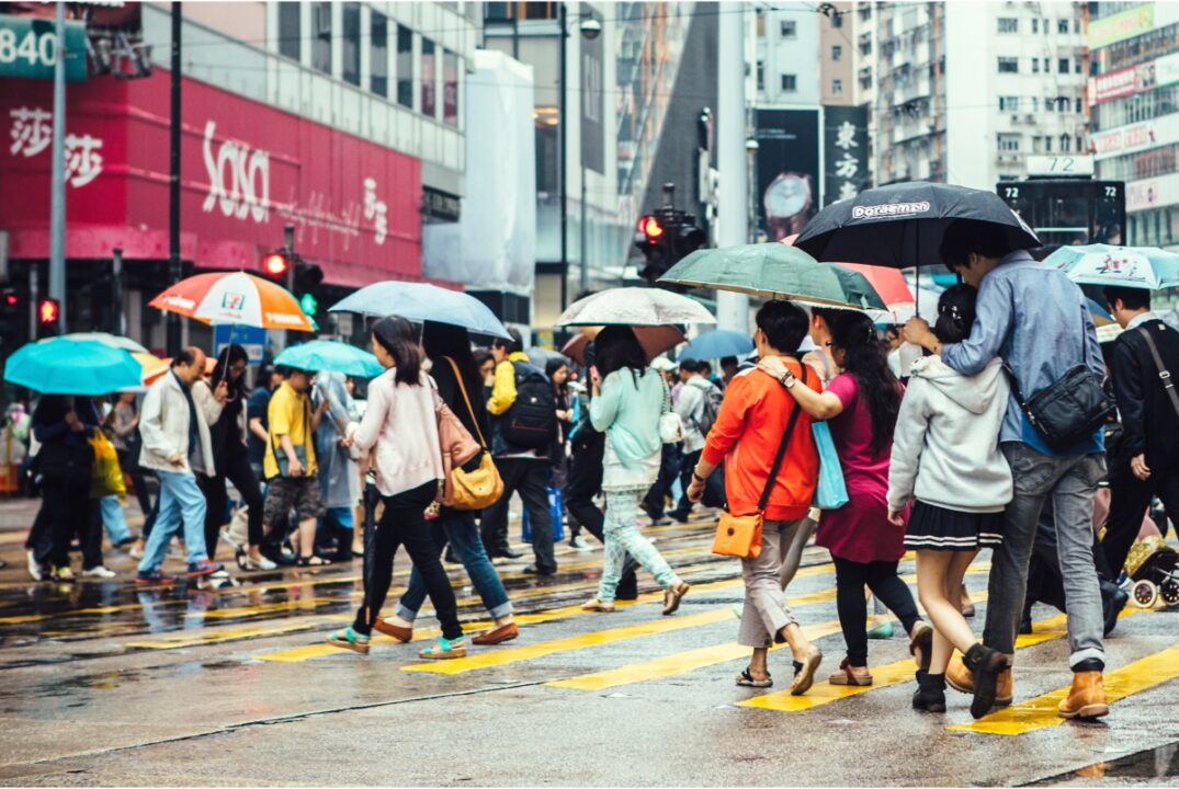 Crowds of people cross a busy Hong Kong street. They are carrying umbrellas and the street is wet.