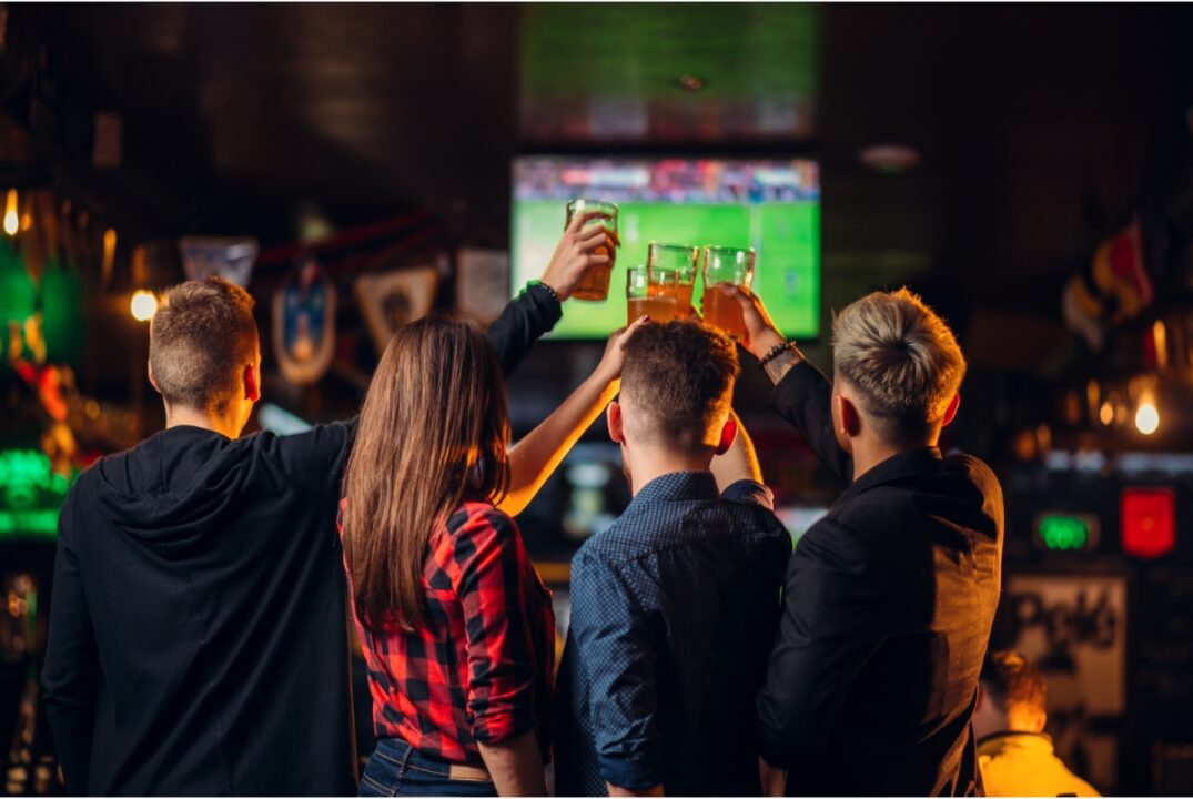 a group of friends in a sports bar raise their glasses in front of a tv showing a football match