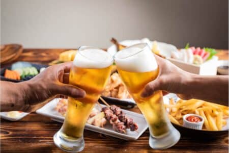 Hong Kong To Give Tourists 1 Million Free Vouchers In November To Use at Restaurants & Bars two people clink their beeg mugs togther in front of a table full of food