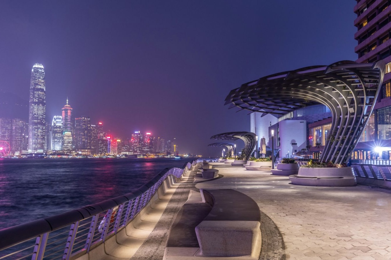 a view of the avenue of stars in hong kong. the promenade is on the right and victoria harbour is on the left, with a view of hong kong island in the background.
