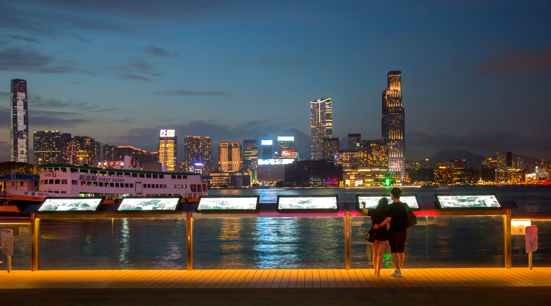 a couple stands in front of a board at the wan chai promenade over looking victoria harbour in hong kong. they can see the kowloon skyline across the harbour as a ferry passes by.