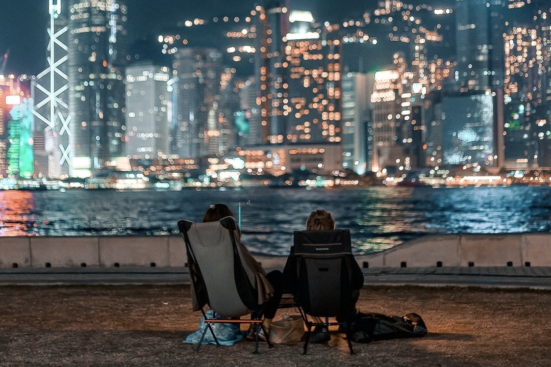 two people sit on foldable seats at art park in west kowloon art park and look at the hong kong island side of the harbour