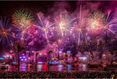 Fireworks light up the sky over Victoria Harbour in Hong Kong. People on the Kowloon side of the harbour watch the display overhead, with the Hong Kong Island skyline in the background.