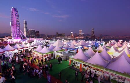 Wine & Dine Is Back at Central Harbourfront from October 23 with 300+ Booths an overhead view of central harbourfront with the hong kong observation wheel in the background. there are food stalls with white tented roofs and people walking around these booths.