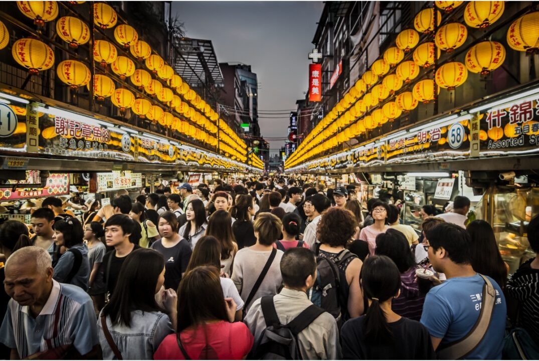 Keelung Night Market in Taiwan. It is crowded with people. There are rows of yellow lanterns suspended above the stalls that are on either side of the people.