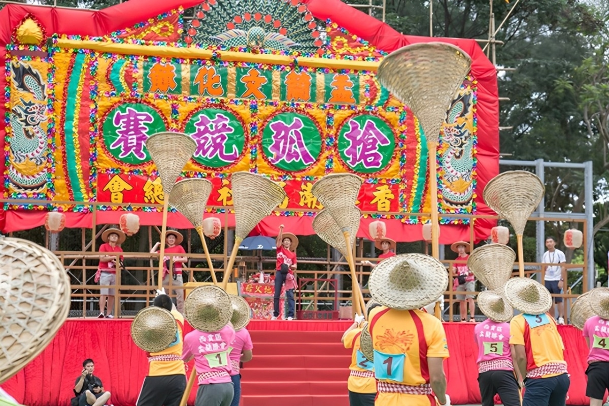 Participants wearing traditional hats and colorful costumes are preparing for a cultural festival performance on a decorated stage featuring vibrant Chinese characters and imagery.