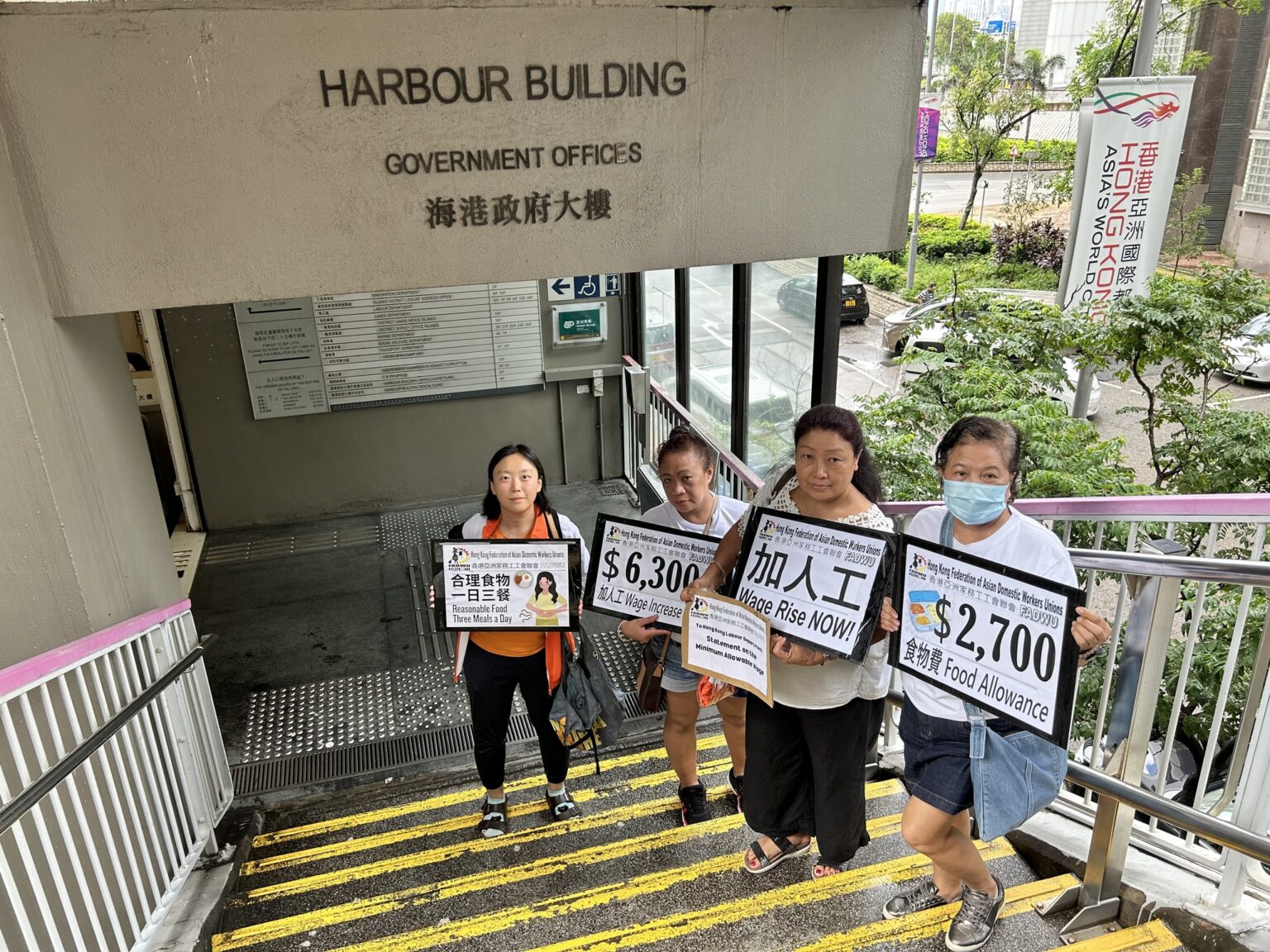 Four domestic helpers hold signs demanding wage increases and better conditions outside the Hong Kong Harbour Building, highlighting issues related to migrant worker wages and labor market conditions in 2025.