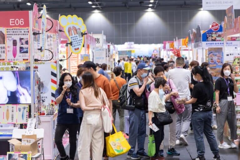 A large crowd of people is browsing various booths at the Hong Kong Brands and Products Expo 2025 in Victoria Park, with some engaging with exhibitors offering products like electronics, abalone deals, and promotional discounts, while many attendees wear masks and carry shopping bags.